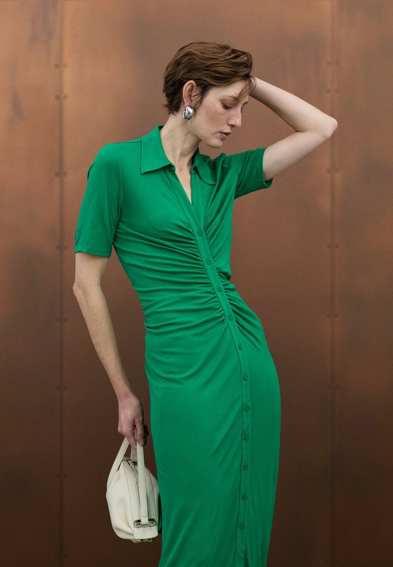 Vestido ajustado verde con mangas cortas, cierre frontal de botones y detalles fruncidos. Combinado con un bolso blanco. Fondo en tonos cálidos de cobre.