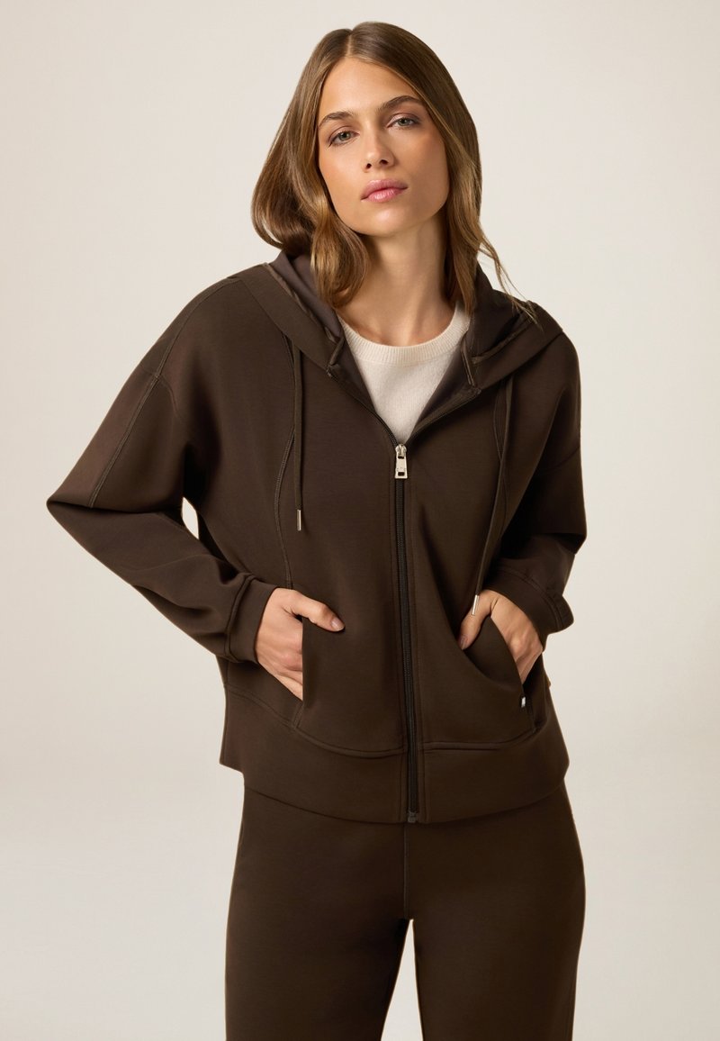 Felpa chiusa marrone realizzata in materiale morbido, con cappuccio, due tasche frontali e una vestibilità rilassata, abbinata a pantaloni coordinati.