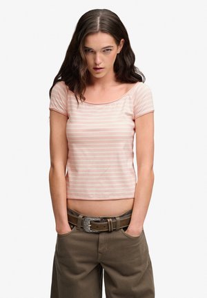 Ung kvinde med langt brunt hår iført en lys pink striped crop top, brune bukser og et dekorativt bælte, hænder i lommerne, hvid baggrund.