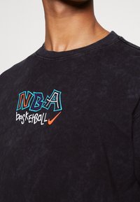Sweatshirt en coton noir avec logo NBA brodé multicolore et texte "basketball", avec le swoosh Nike en orange, design à col rond.