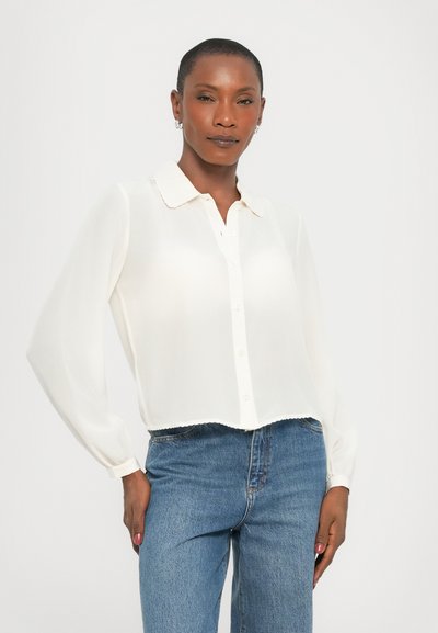 White, long-sleeved blouse with a collar and button closure, features a scalloped hem and sheer material. Worn with high-waisted blue jeans.