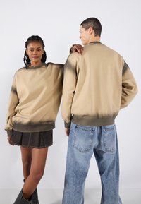 Beige sweatshirt met donkere randen, voorzien van een ronde hals en een losse pasvorm, gecombineerd met een zwarte plooienrok en blauwe wijde jeans.