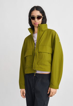 Mujer con chaqueta corta de color verde oliva con cuello alto, pantalones negros con cordón y gafas de sol marrón claro sobre fondo blanco.