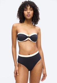 Γυναίκα με σγουρά μαλλιά που φοράει μαύρο μπαντό μπικίνι με λευκή επένδυση και μικρή ξύλινη διακόσμηση στο κέντρο, στέκεται μπροστά σε απλό φόντο.