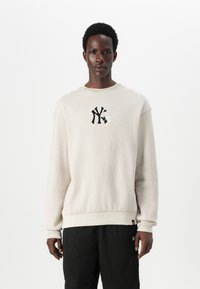 Lys beige ribbestrikket sweatshirt med rund hals, med en svart brodert logo på brystet. Mansjetter og kant har ensfarget farge.