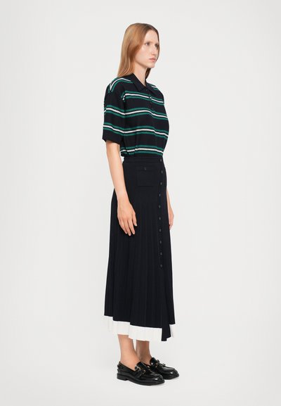 Zwart gestreepte polo shirt met groene en witte strepen, gecombineerd met een zwarte geplooide midi rok met zijknopen en een witte zoom. Zwarte schoenen.