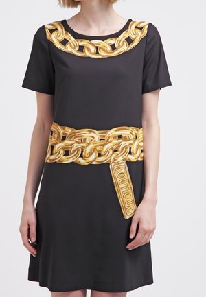 Zwarte jurk met grote geprinte gouden kettingontwerpen rond de halslijn en taille, met een kettinglabel met de tekst "BOUTIQUE."