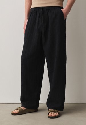 Persona che indossa pantaloni neri larghi e lavorati e sandali aperti color verde oliva, in piedi con le mani nelle tasche su pavimento grigio.