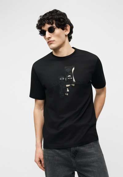 Άνδρας με σγουρά μαλλιά και γυαλιά ηλίου, φορώντας μαύρο μπλουζάκι με αφηρημένα πρόσωπα και σκούρο γκρι τζιν, σε απλό φόντο.