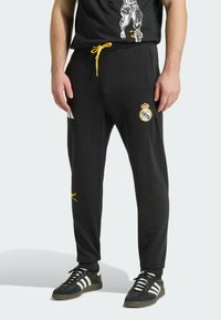 Pantalones deportivos negros con cordón amarillo, que presentan un logo del Real Madrid y un pequeño gráfico de Puma. Hechos de tela suave y elástica.