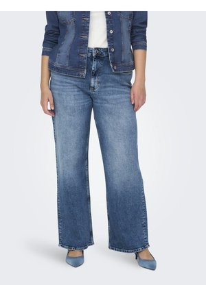 Vrouw in wijde blauwe denim jeans, blauwe denim jacket, witte top en blauwe puntige hakken, staande tegen een effen achtergrond.