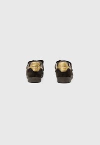 Un paio di sneakers nere in suede con dettagli in oro sul tallone, viste da dietro su uno sfondo grigio chiaro e semplice.
