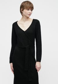 Kvinne med kort hår iført en langermet svart omslagskjole med knytbelte, ser til siden mot en enkel lys bakgrunn.