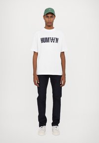 White T-shirt with "HUMAN" print, dark blue jeans, white sneakers, and a green cap. The outfit features a casual, minimalist design.