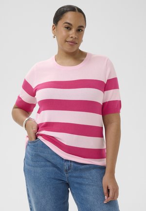 Žena s tmavými vlasmi v krátkom pruhovanom tričku ružovej a tmavoružovej farby a modrých džínsoch, pózujúca s jednou rukou v vrecku, na jednoduchom pozadí.