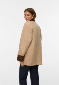 Chaqueta beige con puños de pana marrón, diseño oversize y textura suave. Modelo de perfil, luciendo jeans oscuros.