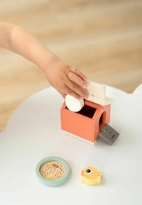 Cutie de jucărie din lemn, de culoare corai, cu un capac alb, care conține o piesă rotundă albă. Un pui de găină galben și un disc cu model se află în apropiere, pe o suprafață albă.