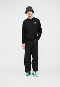 Cămașă neagră cu mânecă lungă, având un grafic pe față, asortată cu pantaloni negri largi și teniși verzi. Pălărie tip bucket de culoare închisă.