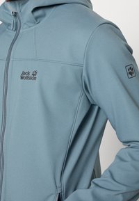 Giacca in pile azzurro chiaro con chiusura lampo frontale, cappuccio e logo ben visibile. Caratterizzata da una texture liscia e dettagli cuciti sulle maniche.