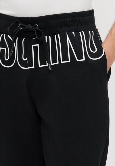 Melni kokvilnas sporta bikses ar aukliņu viduklī un lielu baltu logo tekstu gar sāniem. Materiāls ir mīksts ar gludu tekstūru.