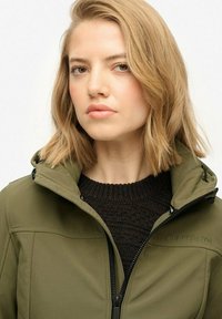 Młoda kobieta z krótkimi blond włosami, nosząca oliwkowo-zieloną kurtkę na zamek nad czarnym swetrem, patrzy wprost z neutralnym wyrazem twarzy.