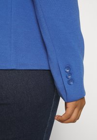 Niebieska dzianinowa kurtka o fakturowanej tkaninie, z trzema pasującymi guzikami na mankiecie oraz prostym, dopasowanym kroju. Widoczny ciemny dżins.