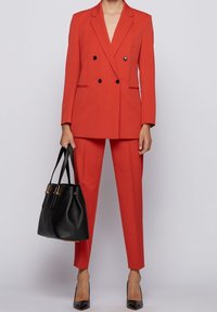 Veste blazer rouge croisée et pantalon slim assorti en tissu lisse. Sac à main noir avec accents dorés et escarpins noirs pointus.