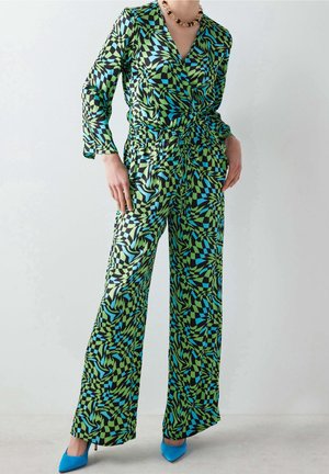 Personne portant une combinaison à manches longues à motifs géométriques verts, noirs et bleus, avec des jambes larges et des escarpins bleus pointus.