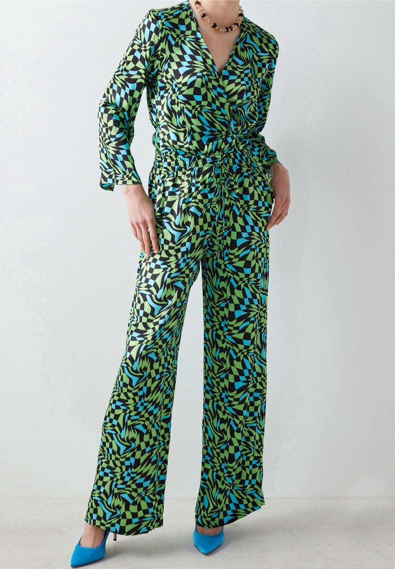 Personne portant une combinaison à manches longues à motifs géométriques verts, noirs et bleus, avec des jambes larges et des escarpins bleus pointus.