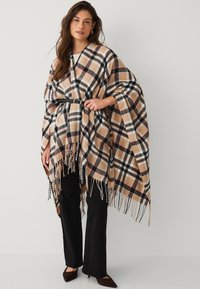Plaid poncho with a beige, black, and red color scheme. Finished with fringes and a black belt at the waist, worn over black pants.