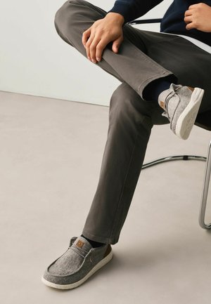 Osoba siedząca na krześle, skrzyżowane nogi, ubrana w szare, codzienne buty, ciemne spodnie i ciemną bluzkę z długim rękawem, na jednolitej podłodze.