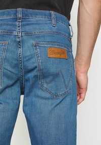 Jeans in denim blu con una toppa in pelle Wrangler sulla tasca posteriore, caratterizzati da cuciture a contrasto e una vestibilità classica. Texture liscia.