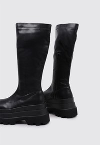 Botas negras de cuero hasta la rodilla con un diseño elegante, que presentan una suela de plataforma gruesa y una cremallera lateral para facilitar su uso.