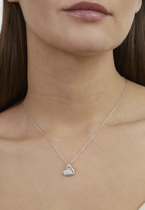 Collier avec pendentif cœur en argent sur une chaîne délicate, présentant une surface polie et un design subtilement fendu, reposant sur un cou nu.