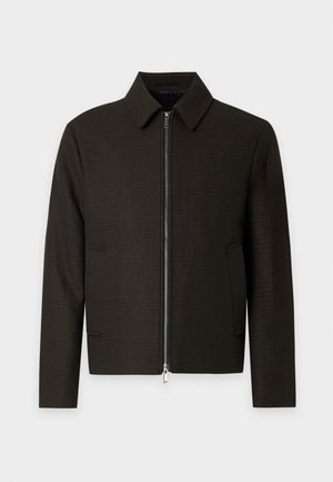 Mand iført en mørk lynlåstrøje, blå jeans og brune sneakers, der står mod en ensfarvet lys baggrund.