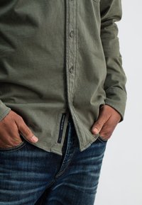 Olijfgroen overhemd met knopen, gemaakt van een gestructureerde stof, met knopen aan de voorkant en een label aan de zoom. Draagt donkere blauwe spijkerbroek, handen in de zakken.