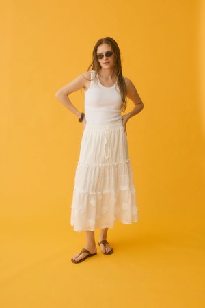 Vrouw met een wit mouwloos topje en een gelaagde witte rok, zonnebril en sandalen, staand tegen een oranje achtergrond met handen in de zij.