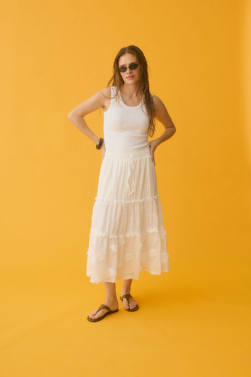 Femme portant un top blanc sans manches et une jupe blanche à volants, avec des lunettes de soleil et des sandales, debout les mains sur les hanches devant un fond orange.