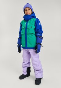 Niebieska i zielona kurtka narciarska z dwoma przednimi kieszeniami, fioletowy beanie, lawendowe spodnie i czarne buty śniegowe. W zestawie rękawice i wysoki kołnierz.