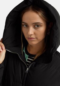 Černá péřová bunda s velkou kapucí, s předním zipem a prošívanou texturou. Pod ní je částečně vidět pruhované tričko.