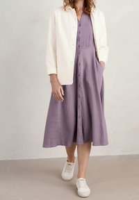 Kvinna iklädd en ljuslila midiklännings med knappar framtill och fickor, krämfärgad jacka och vita snörskor, stående på en beige golv.