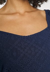 Vestido azul-marinho com decote quadrado, apresentando detalhes de ilhós estampados. O tecido é leve com um acabamento texturizado.