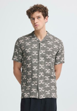 Młody mężczyzna z kręconymi włosami, ubrany w krótką koszulę na guziki z powtarzającym się abstrakcyjnym białym wzorem na szarym materiale.