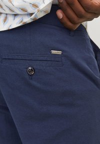 Laivastonsiniset housut, jotka on valmistettu kevyestä kankaasta. Niissä on napillinen kiinnitys ja pieni metallinen logotunnus takataskussa.