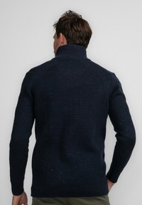 Marineblau gestrickter Pullover mit hohem Kragen, strukturiertem Muster und gerippten Bündchen sowie Saum. Die Rückansicht zeigt kunstvolle Nähdetails.