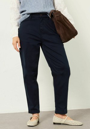 Frau trägt marineblaue Hose, hellblauen Strickweste über weißer Spitzenbluse, trägt braune Wildledertasche und cremefarbene flache Schuhe.