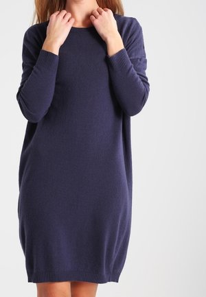 Femme portant une robe-pull bleu marine à manches longues, longueur genoux, tenant l'encolure des deux mains.