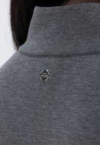 Sweat-shirt gris avec un col montant et un badge rond épinglé orné de pierres décoratives claires près du col. Texture de tissu lisse.