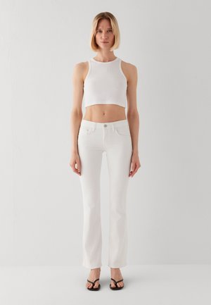 Frau mit kurzen blonden Haaren steht und trägt ein weißes ärmelloses Croptop, weiße Jeans und schwarze Sandalen mit offenen Zehen vor einem schlichten weißen Hintergrund.
