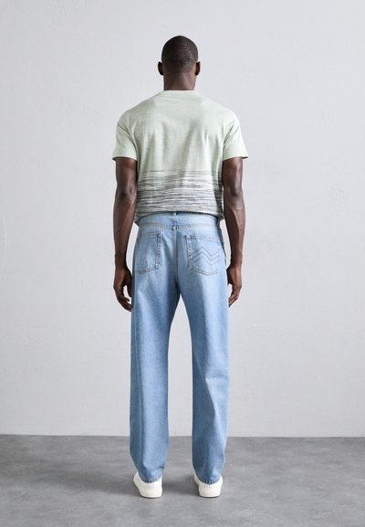 Light green t-shirt with a dark striped pattern, paired with loose blue denim jeans and white sneakers, showcased from the back.